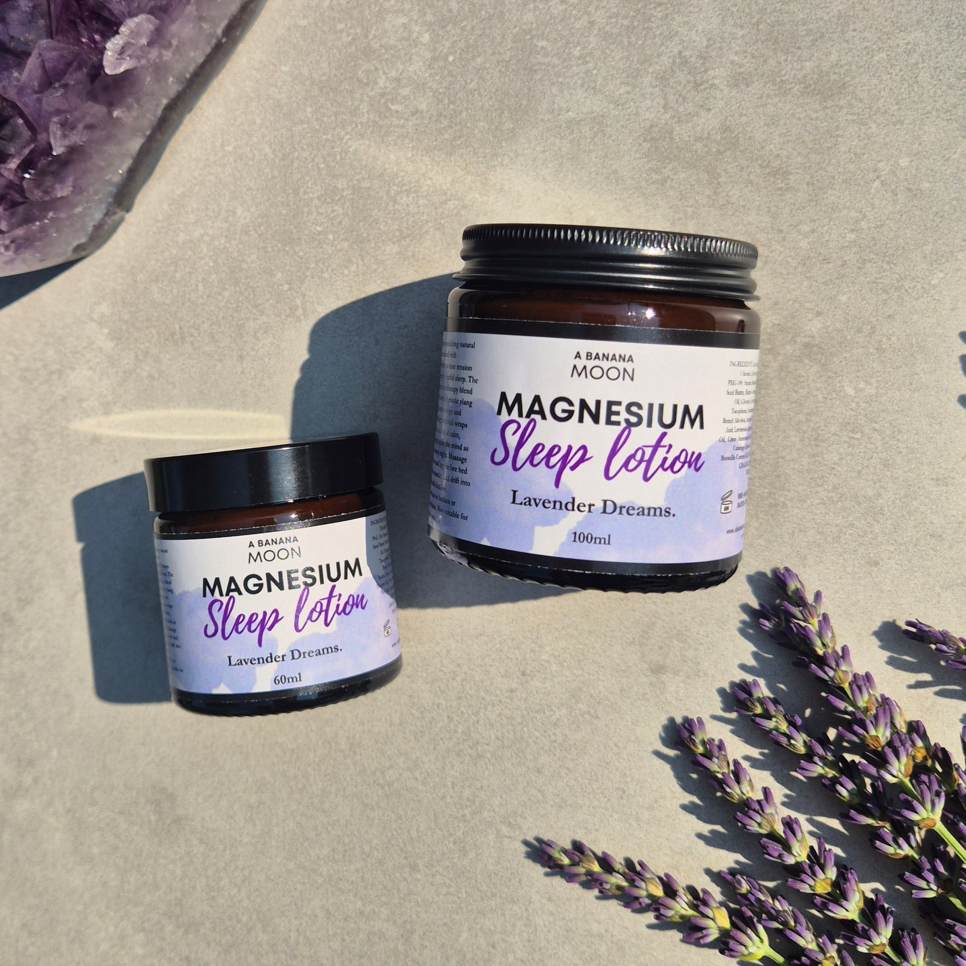 Two jars of A Banana Moon Magnesium Sleep Lotion on a textured surface with lavender flowers.